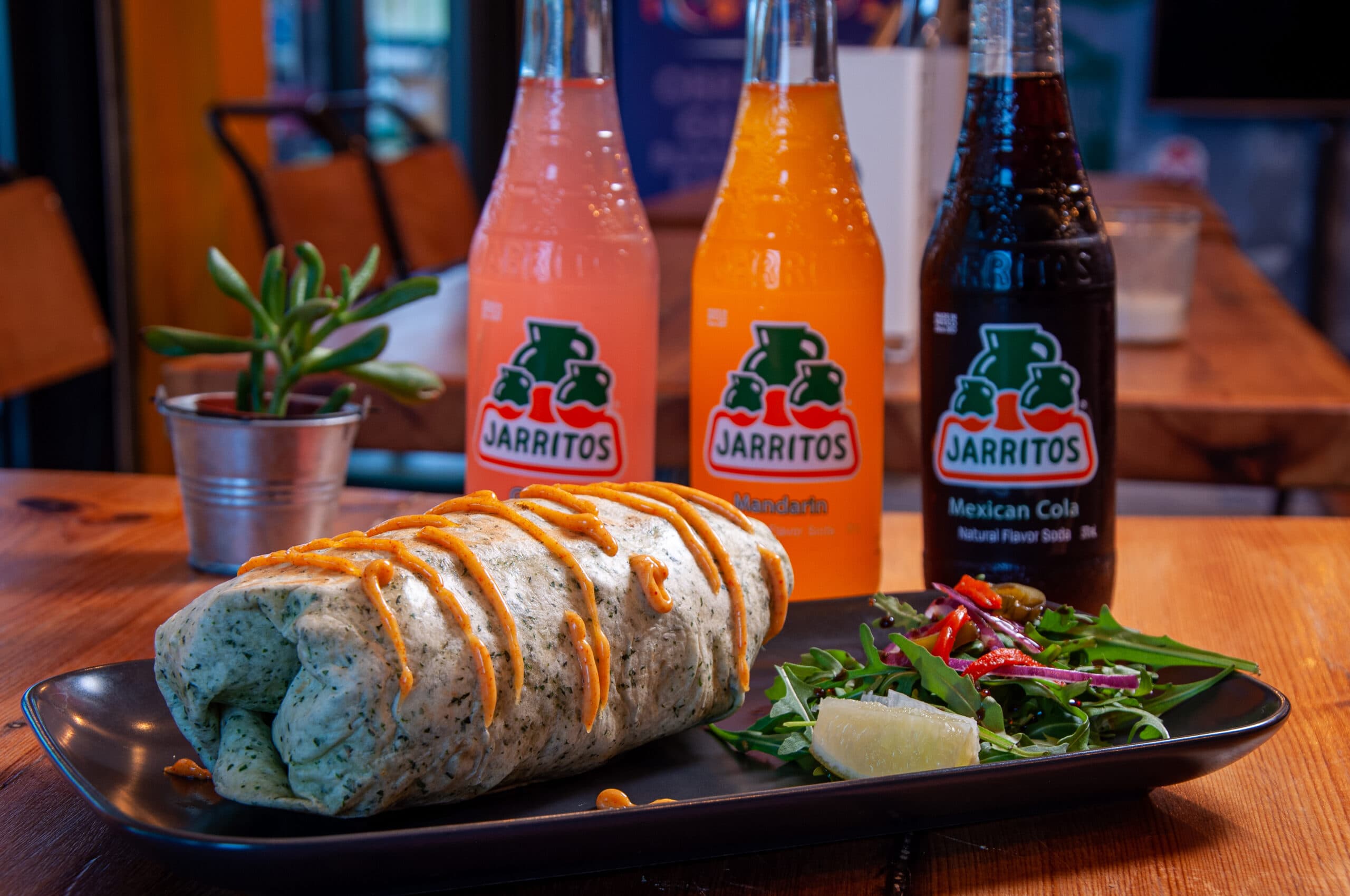A View Tube burrito with chipotle drizzle beside three colourful Jarritos Mexican sodas on the warm wooden bar.